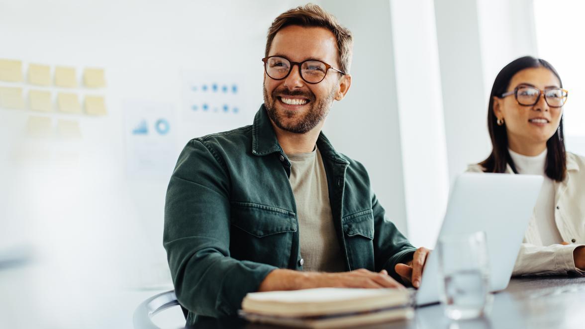 Two happy people working in a light modern office with notes on a whiteboard behind them and a laptop