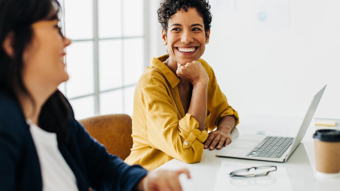 Two people in a light modern office laughing in a meeting with a laptop