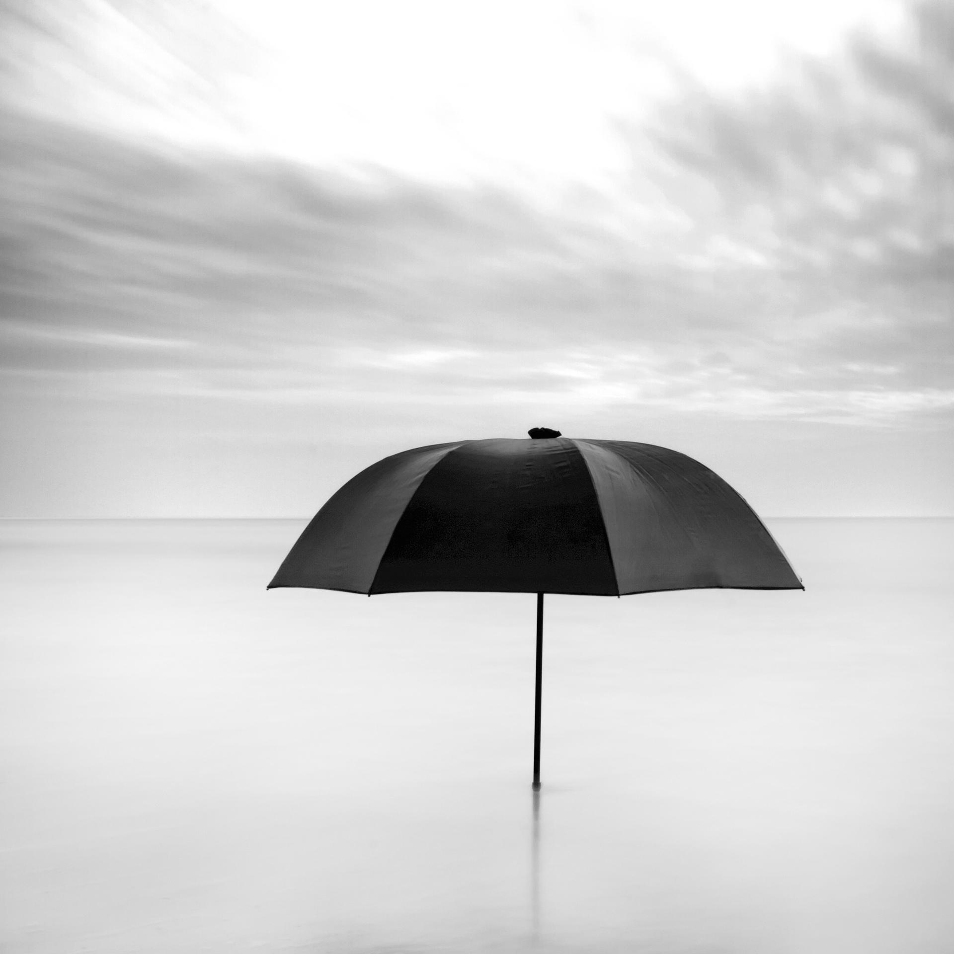 A black and white image of a striped umbrella emerging from some water with a bright and cloudy sky behind it