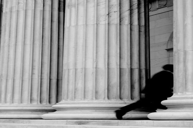 A figure in a suit and gown enters between stone columns of a building