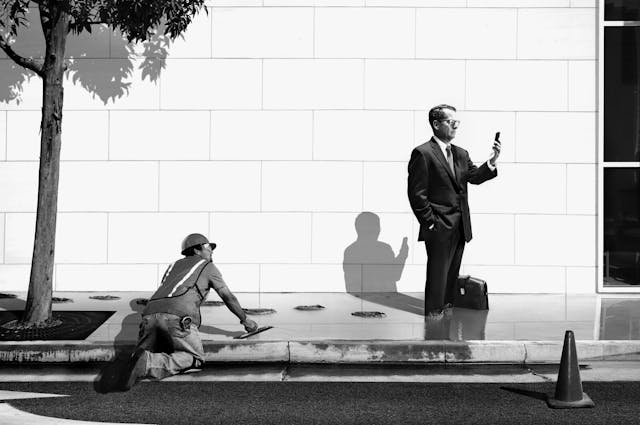Man in suit looking at his phone walks through wet concrete paving while the man on his hands and knees working on smoothing the concrete looks up at him